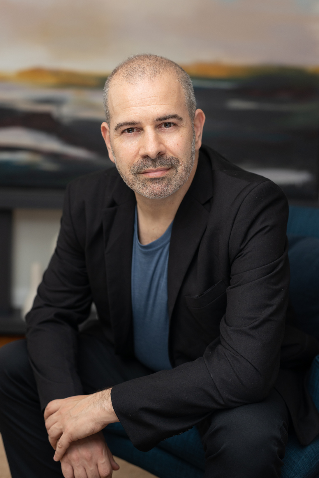 Half body image of Jonathan Marion, seated, facing the camera, wearing a black suit with a blue shirt, with a multicolored painting featuring golden and blue hues as the background.